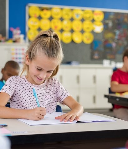 Portrait of happy girl sitting at school desk studying in classroom. Smiling schoolgirl doing classwork with classmates in background. Clever female student taking notes on notebook at primary school.